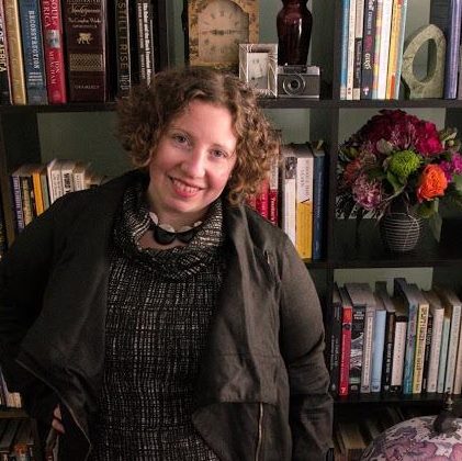 Cara Berg Powers a white woman standing in front of a book case with flowers on her right side. She is wearing a gray sweater and gray jacket